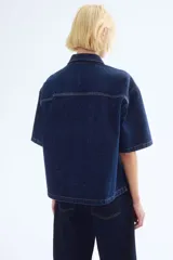 Camisa azul de denim, corte amplio, con cuello clásico, abotonadura frontal, manga corta, bolsillo de parche en el pecho y canesú en la espalda.