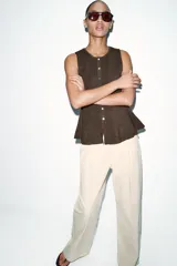 Top de punto acanalado en color beige, sin mangas y con cuello redondo. Presenta un cierre frontal con botones pequeños y un bajo con acabado en peplum que aporta volumen.