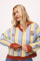 Sweater de punto con cuello polo, a rayas verticales celestes y amarillas, con puños, cuello y ruedo en color marrón con detalles en rojo. Presenta un parche bordado con el logo de la marca en el pecho y un bordado con una letra B en la manga.