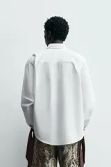 Camisa blanca de algodón popelín, corte relajado, con cuello solapa y pechera frontal plisada. Las mangas terminan en puños ajustables con lazos largos estampados en color marrón con patrón circular.