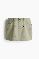 Pollera corta de denim color verde oliva, estilo cinco bolsillos, con cierre y botón frontal. Presenta apliques de tachas metálicas doradas y plateadas en forma de estrella o brújula distribuidas en el frente.