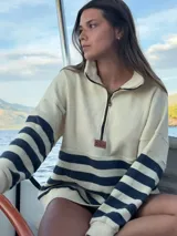 Buzo estilo marinero color crudo con franjas horizontales azul marino, confeccionado en 100% lana. Presenta cuello alto con cierre medio, puños y cintura ajustados.