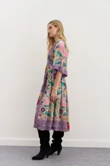 Vestido largo de viscosa con mangas al codo y falda amplia. Presenta un estampado floral vibrante sobre fondo beige, con detalles y bordes en tonos morados y patrones geométricos. Tiene escote en V y cierre trasero.