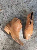 Botas de cuero color suela, de estilo texano, con caña baja, puntera fina y detalles de costuras decorativas.