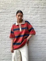 Remerón a rayas horizontales color marrón, azul y rojo, con cuello tipo polo color beige y mangas cortas con puños tejidos.