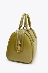 Bolso formato mini bowling color verde oliva con detalles metálicos dorados en las asas. Tiene doble asa de mano y correa bandolera extraíble. Incluye bolsillo interior con cremallera y cierre principal con cremallera.