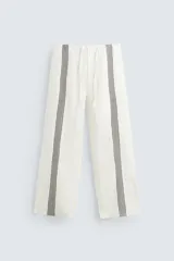 Pantalón de lino color crudo, de corte recto y tiro alto, con cintura elástica ajustable con cordón. Presenta bolsillos laterales y traseros, y bandas verticales negras a lo largo de las piernas.