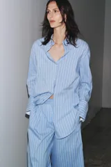 Camisa celeste con rayas verticales blancas, de corte amplio, cuello clásico, manga larga con puños y cierre frontal con botones.