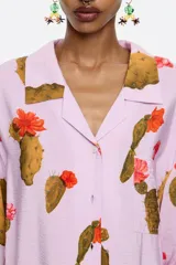 Camisa de viscosa color lila con estampado de cactus y flores rojas, corte recto, cuello con solapas, manga larga recta, cierre frontal con botones y bolsillos de plastrón.