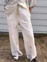 Pantalón palazzo blanco de tiro alto, corte ancho y holgado, con bordados en hilo dorado o beige claro. Presenta un cordón trenzado con borlas en la cintura.