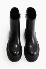 Botas negras de caña media confeccionadas en cuero sintético, con puntera redondeada y cierre lateral con cremallera. Presentan suela track de goma.