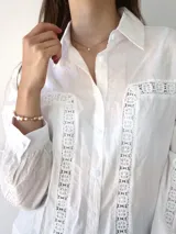 Camisa blanca de mangas largas con detalles de broderie en el frente, mangas y espalda. Corte recto, cuello clásico y cierre frontal con botones.