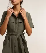 Vestido midi color verde militar, estilo camisero, con cuello clásico, mangas cortas, abotonadura frontal completa, bolsillos utilitarios en el pecho, lazo en la cintura y falda acampanada.