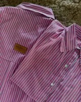 Camisa de manga larga con cuello y botones frontales, confeccionada en tela satinada a rayas verticales fucsia, blancas y negras. Presenta un bolsillo en el pecho y un parche de cuero con el logo de la marca.