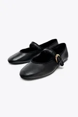 Zapato plano tipo bailarina color negro, confeccionado en piel, con tira sobre el empeine y hebilla metálica dorada ajustable. Tiene punta redonda y suela de 1.5 cm de altura.