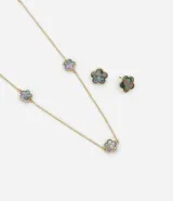 Conjunto de bisutería compuesto por un collar largo con tres dijes de flores y caravanas tipo botón con forma de flor. Las piezas cuentan con un acabado iridiscente de nácar y una estructura de metal con baño dorado.