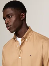 Camisa color beige de corte regular, confeccionada en algodón elástico. Cuenta con cuello abotonado, mangas largas con puños abotonados y logo de Tommy Hilfiger bordado en el pecho.