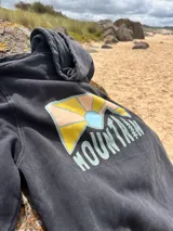 Buzo hoodie negro con capucha y cordones ajustables. Estampado en la espalda con un diseño de sol naciente y la palabra "Mountain" en letras color celeste.