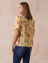 Blusa amarilla estampada con flores rosadas, de gasa, con cuello a la base, escote en V plisado y lazo de ajuste. Delantera con detalles en puntilla y mangas 3/4 con puño fruncido.