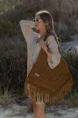 Bolso grande tejido a crochet con trapillo color marrón, con flecos en la parte inferior y etiqueta de cuero con el logo de la marca.