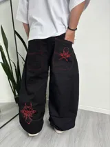 Pantalón baggy negro con costuras rojas contrastantes y bordados rojos de calavera y cruz en el bolsillo trasero y la pierna izquierda.
