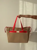 Bolso tipo tote de color marrón con detalles en rojo. Tiene cierre superior, monedero, bolsillo interno y externo con cierre, compartimentos internos y opción de correa larga.