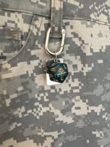 Llavero metálico plateado con mosquetón, que incluye varios dijes: una placa rectangular con el As de Picas, un dado poliédrico verde oscuro y dorado, una estrella de cuatro puntas, y un dije pequeño con forma de corazón que dice "made with love".