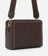 Cartera transversal de material sintético color marrón oscuro, de forma rectangular y estructura rígida. Presenta pespuntes decorativos en color blanco en los bordes frontales y cierres metálicos dorados. Incluye un asa ancha desmontable con diseño de rayas beige y marrón.