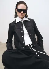 Chaqueta corta de cuero negro con cuello camisero, cierre frontal con botones a presión plateados, bolsillos laterales y puños abotonados.