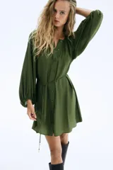Vestido corto color verde oliva, de corte holgado, con cuello redondo fruncido, mangas globo largas con puños elásticos y botonadura frontal. Incluye cinturón fino para atar.