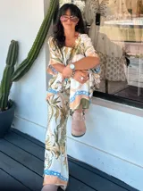 Pantalón ancho de satén con cintura elástica y estampado de motivos florales y cadenas en tonos marrones, beige y dorado sobre fondo blanco. Presenta un borde inferior con detalles en color turquesa.