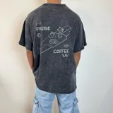 Remera negra con efecto gastado, corte oversize y estampa en la espalda con la frase 'Pause & Coffee UW' y dibujo de una mesa con café y medialunas.