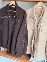 Chaqueta color beige con bordado calado, cuello camisero, cierre frontal con botones, mangas largas con puños abotonados, bolsillos de parche en el pecho y ruedo desflecado.