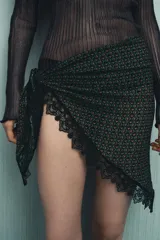 Pañuelo tipo pareo estampado con un patrón geométrico en tonos verde, naranja y negro. Presenta un borde de encaje negro a contraste.