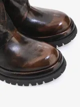 Bota corta de cuero bovino con efecto envejecido y desgastado en tonos marrón oscuro y negro. Presenta un diseño robusto con caña arrugada, cierre lateral diagonal con tirador metálico dorado y suela track gruesa.