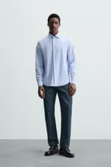 Camisa celeste a rayas verticales blancas, de corte recto, con cuello italiano abierto, mangas largas con puños abotonados y cierre frontal con botones.
