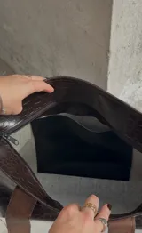 Bolso tote negro con textura símil cuero de cocodrilo, bolsillo marrón con tachas plateadas en el frente y asas de cuero marrón.