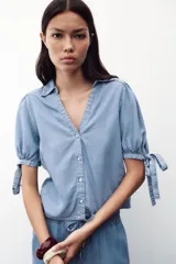 Camisa celeste de denim con cuello solapa, escote en pico, mangas abullonadas con lazos y cierre frontal con botones.