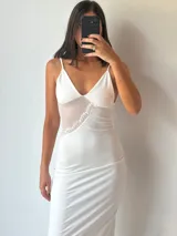 Vestido largo blanco, ajustado al cuerpo, con escote pronunciado en V y tirantes finos. Presenta un detalle de encaje o puntilla en el lateral del busto.