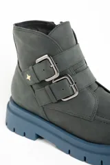 Bota corta de cuero color verde oscuro, con suela track gruesa de color celeste. Presenta dos correas ajustables con hebillas metálicas plateadas y un pequeño dije de estrella dorada en el lateral. Tiene cierre de cremallera en el interior.