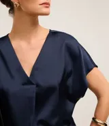 Blusa color café confeccionada en poliéster y elastano, con acabado satinado, escote V y mangas cortas. Presenta pliegues delanteros.