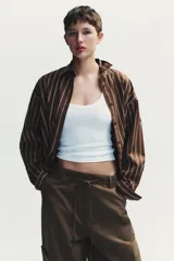 Camisa oversize marrón con rayas verticales blancas, cuello solapa, manga larga con puños y hombros caídos. Tiene un bolsillo de plastrón en el delantero, pinzas en la espalda y cierre frontal con botones.