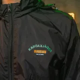 Campera negra tipo windbreaker con capucha, cierre frontal y puños elásticos. Logo de Kaboa en el pecho y estampado con el logo de Jägermeister en la manga.