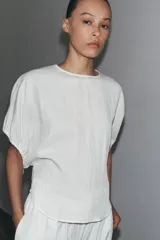 Blusa blanca de lyocell con cuello redondo, manga corta abullonada ajustable con cordones, bajo redondeado y cierre en la espalda con abertura y botón.