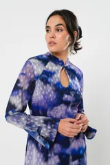 Vestido largo con estampado abstracto en tonos azules, violetas y grises. Tiene cuello mao con abertura en forma de gota, mangas largas acampanadas con detalle de botón en los puños, espalda abierta, tajo frontal y flecos en el ruedo.