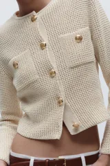 Chaqueta color crema de tejido de punto con cuello redondo y manga larga. Tiene bolsillos delanteros de plastrón y cierre frontal con botones dorados.