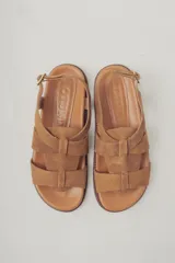 Sandalia plana de gamuza color marrón oscuro, con tiras cruzadas en la parte delantera y una tira fina ajustable con hebilla metálica en el tobillo. Posee plantilla de cuero y suela baja.