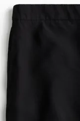 Pantalón negro de corte ancho y tiro alto, con elástico en la parte posterior de la cintura, cierre oculto con corchete en un lado, bolsillos laterales discretos y pliegues adelante y atrás.