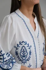 Camisa blanca de manga larga con elástico en puños, cuello mao con botones en la parte delantera y bordados florales azules en el frente y las mangas.