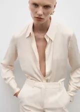 Camisa color crema de tejido fluido con textura y acabado satinado. Tiene cuello clásico, cierre de botones frontal y mangas largas con puños abotonados.